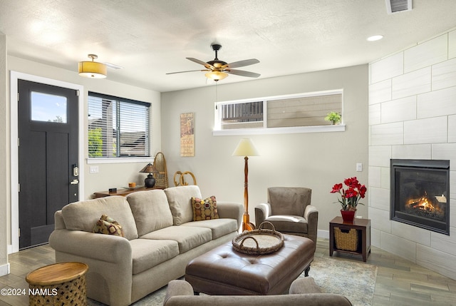 living room featuring ceiling fan, a textured ceiling, and a tiled fireplace