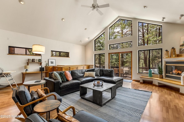 living room featuring high vaulted ceiling, a glass covered fireplace, a ceiling fan, baseboards, and wood finished floors