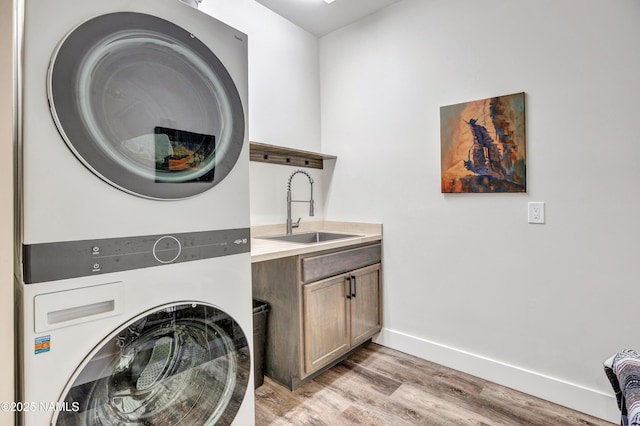 clothes washing area featuring a sink, stacked washer / drying machine, baseboards, light wood-type flooring, and cabinet space