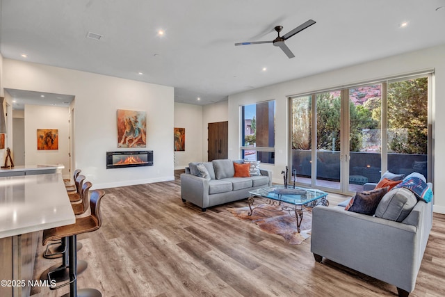 living room featuring light wood-style flooring, ceiling fan, baseboards, a glass covered fireplace, and recessed lighting