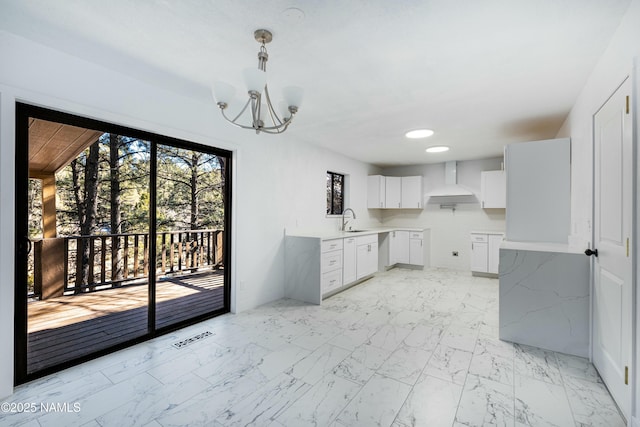 kitchen with wall chimney exhaust hood, sink, an inviting chandelier, hanging light fixtures, and white cabinets