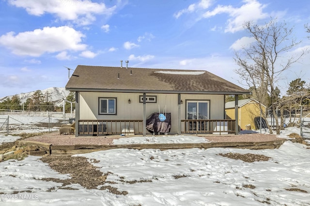 snow covered property with a mountain view, roof with shingles, and fence