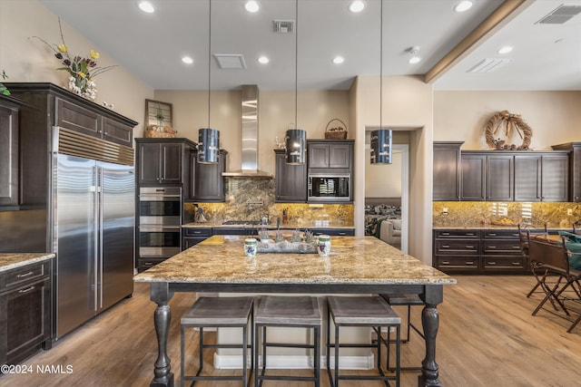 kitchen featuring a large island with sink, wall chimney exhaust hood, decorative light fixtures, built in appliances, and dark brown cabinets