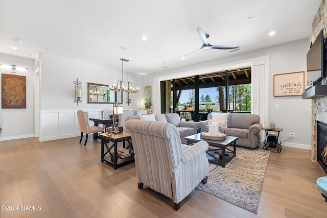 living room with ceiling fan with notable chandelier, a stone fireplace, and light wood-type flooring