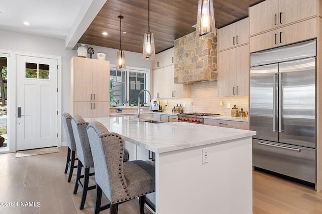 kitchen with light brown cabinets, hanging light fixtures, sink, a kitchen island with sink, and built in fridge