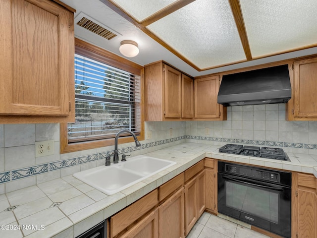 kitchen featuring stainless steel gas stovetop, sink, oven, tile countertops, and wall chimney range hood