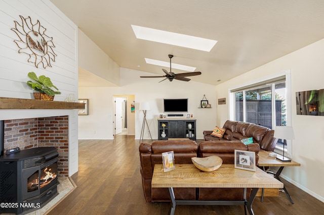 living area with vaulted ceiling with skylight, baseboards, a wood stove, ceiling fan, and wood finished floors