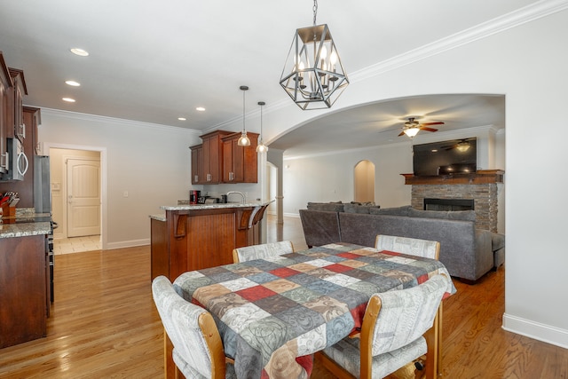 dining room with light hardwood / wood-style flooring, ceiling fan with notable chandelier, sink, a stone fireplace, and ornamental molding