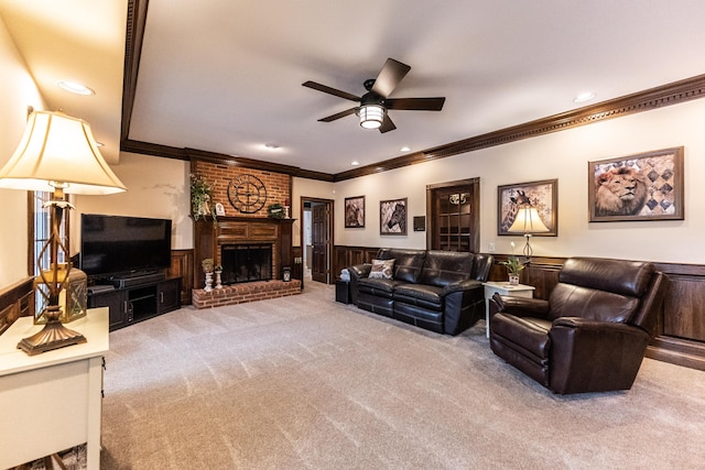 living room with a wainscoted wall, carpet flooring, crown molding, a brick fireplace, and ceiling fan