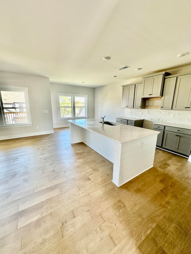 kitchen with gray cabinetry, tasteful backsplash, sink, light wood-type flooring, and a center island with sink