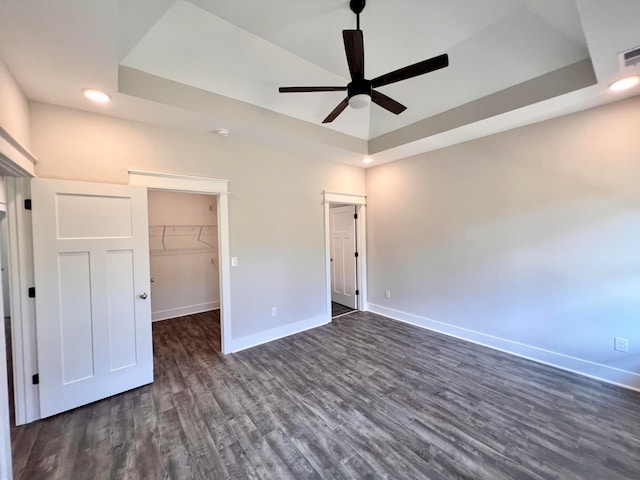 unfurnished bedroom with a walk in closet, dark hardwood / wood-style flooring, a closet, a tray ceiling, and ceiling fan