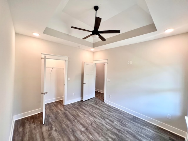 unfurnished bedroom featuring a closet, a walk in closet, a raised ceiling, dark wood-type flooring, and ceiling fan