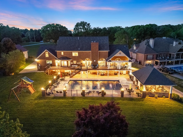 back house at dusk featuring a playground, a swimming pool, a lawn, and a patio