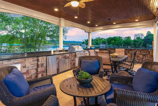 patio terrace at dusk with a water view, an outdoor kitchen, sink, ceiling fan, and grilling area