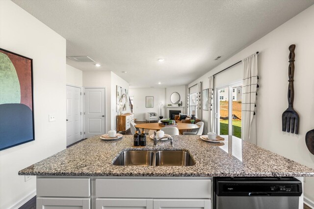 kitchen with stone counters, a textured ceiling, a center island with sink, sink, and stainless steel dishwasher