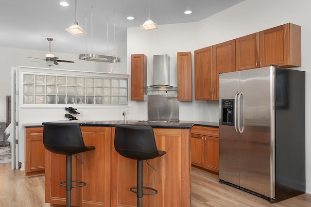 kitchen with light hardwood / wood-style flooring, ceiling fan, stainless steel refrigerator with ice dispenser, wall chimney exhaust hood, and a breakfast bar area