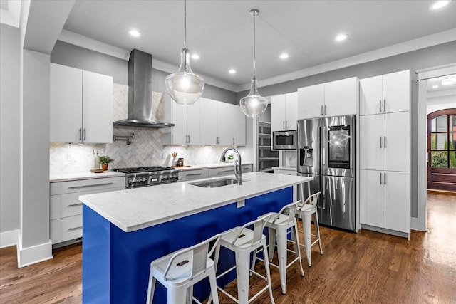 kitchen featuring appliances with stainless steel finishes, a kitchen island with sink, wall chimney range hood, sink, and white cabinets