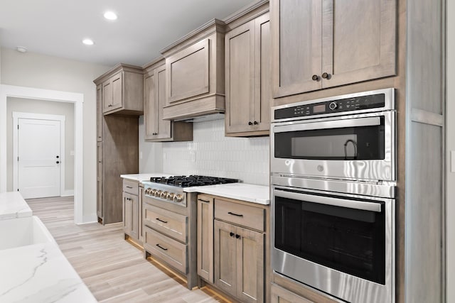 kitchen featuring appliances with stainless steel finishes, backsplash, light stone counters, custom range hood, and light hardwood / wood-style flooring