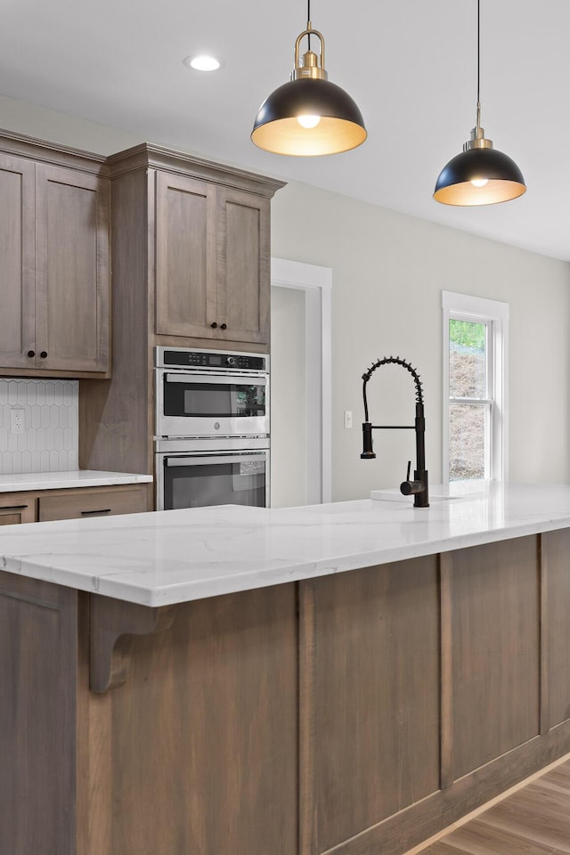 kitchen featuring backsplash, light stone counters, stainless steel double oven, hardwood / wood-style floors, and hanging light fixtures