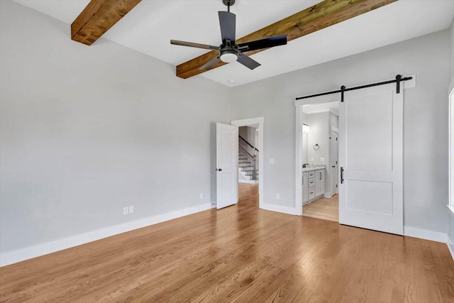 unfurnished bedroom with beamed ceiling, a barn door, light wood-type flooring, ensuite bathroom, and ceiling fan