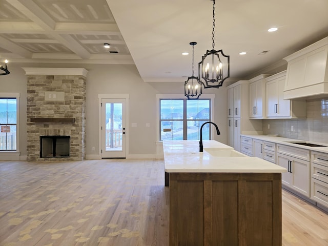 kitchen featuring coffered ceiling, a kitchen island with sink, sink, white cabinets, and decorative light fixtures