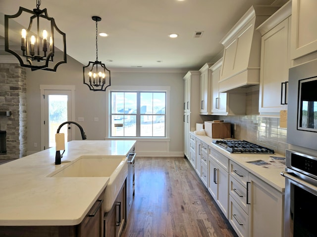 kitchen with pendant lighting, appliances with stainless steel finishes, custom range hood, a center island with sink, and a chandelier