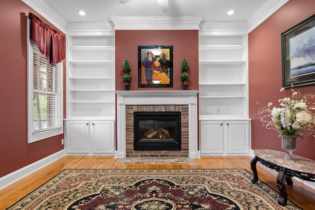 living room featuring a brick fireplace, light hardwood / wood-style flooring, built in shelves, ceiling fan, and ornamental molding