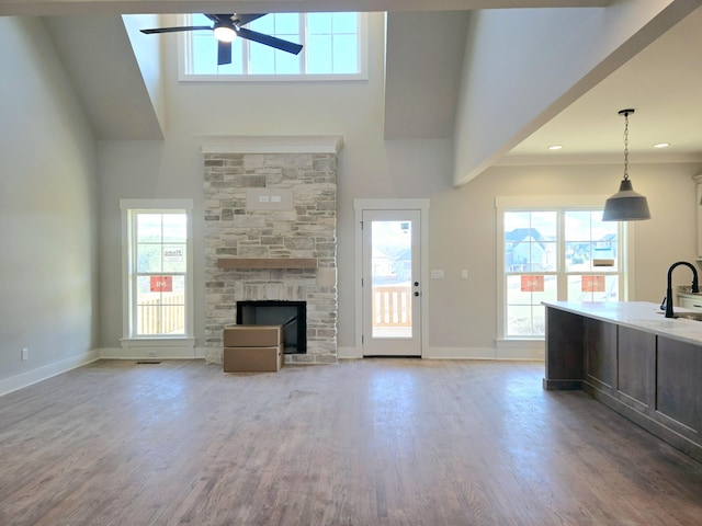 unfurnished living room with sink, ceiling fan, a high ceiling, a stone fireplace, and light wood-type flooring