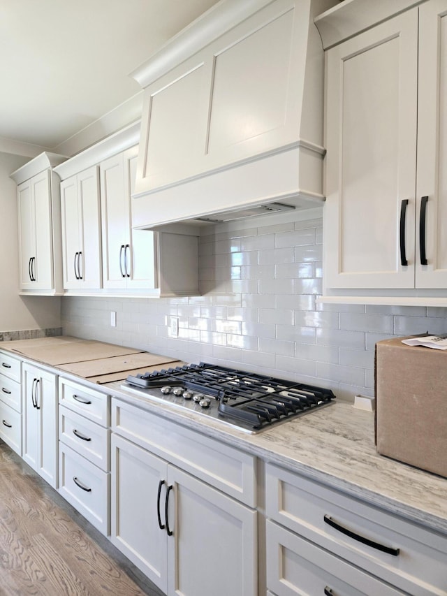 kitchen featuring stainless steel gas cooktop, custom exhaust hood, white cabinetry, tasteful backsplash, and light hardwood / wood-style floors