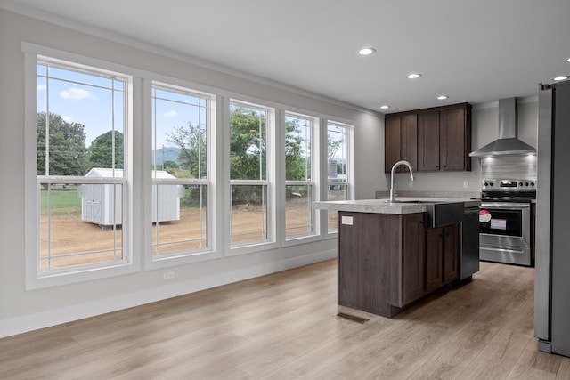 kitchen with dark brown cabinets, a center island with sink, wall chimney range hood, appliances with stainless steel finishes, and light wood-type flooring