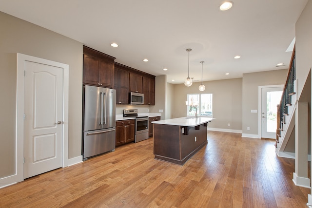 kitchen with appliances with stainless steel finishes, an island with sink, hanging light fixtures, dark brown cabinetry, and light wood-type flooring