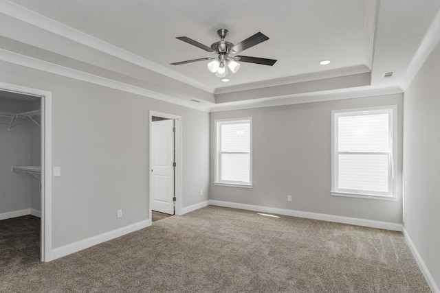 unfurnished bedroom featuring ceiling fan, a raised ceiling, carpet floors, a walk in closet, and ornamental molding