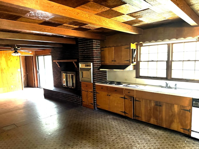 kitchen with beam ceiling, wall oven, brown cabinetry, light countertops, and a brick fireplace