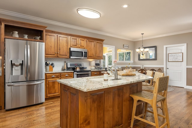 kitchen with light wood-type flooring, appliances with stainless steel finishes, light stone countertops, a notable chandelier, and a center island with sink