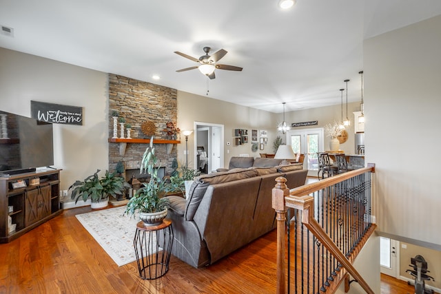 living room with ceiling fan with notable chandelier, wood-type flooring, and a stone fireplace