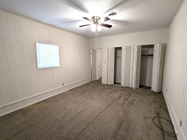 unfurnished bedroom with wooden walls, a textured ceiling, dark carpet, ceiling fan, and multiple closets