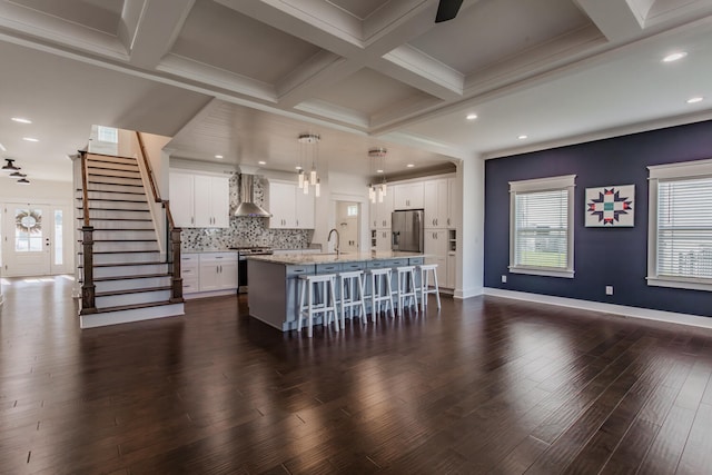 kitchen featuring white cabinetry, decorative light fixtures, stainless steel appliances, an island with sink, and wall chimney range hood