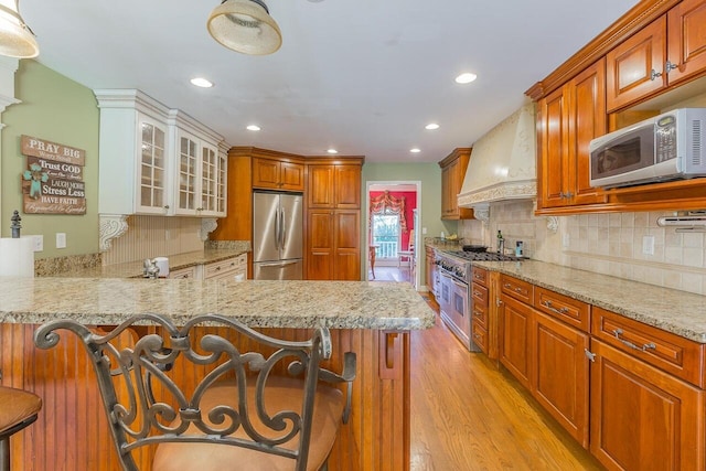 kitchen with light wood-type flooring, stainless steel appliances, premium range hood, kitchen peninsula, and a kitchen bar