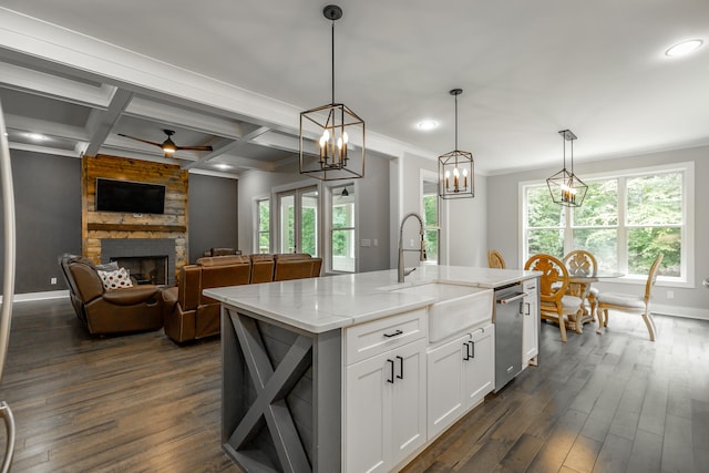 kitchen with dark wood-type flooring, ceiling fan with notable chandelier, a kitchen island with sink, and a stone fireplace