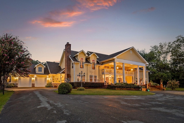 greek revival inspired property with driveway, a chimney, an attached garage, a balcony, and a lawn