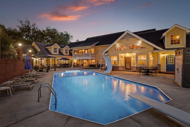 pool at dusk with a water slide, an outdoor pool, a patio, and a diving board