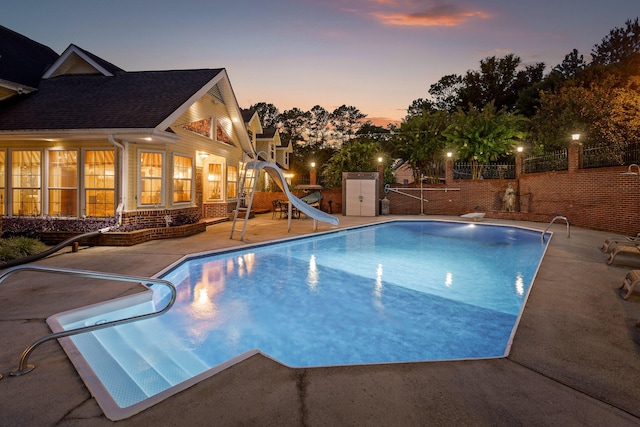 pool at dusk with fence, a water slide, a fenced in pool, a diving board, and a patio area