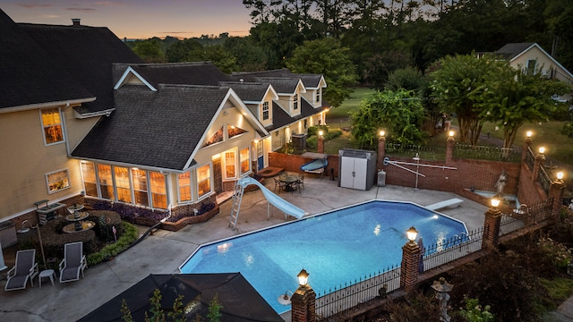 pool at dusk with a water slide, a diving board, and a patio