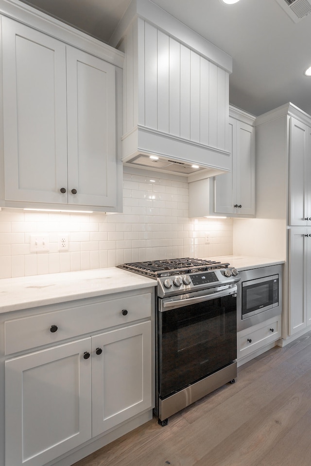 kitchen with tasteful backsplash, white cabinets, custom range hood, light hardwood / wood-style flooring, and appliances with stainless steel finishes