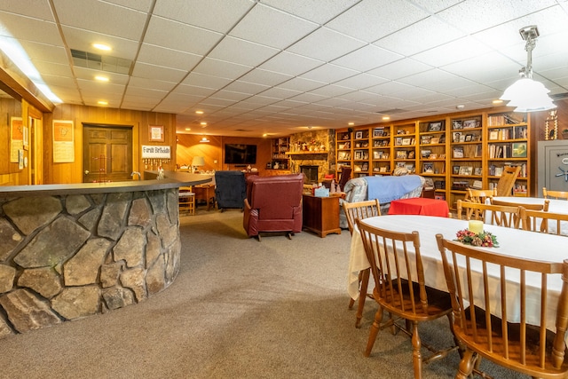 carpeted dining space with wooden walls, a drop ceiling, and a stone fireplace