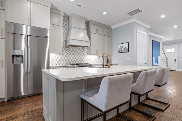 kitchen with a kitchen bar, premium range hood, a kitchen island with sink, sink, and stainless steel built in fridge