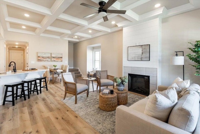 living room with a tiled fireplace, light wood-type flooring, beamed ceiling, coffered ceiling, and ceiling fan