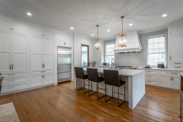 kitchen with custom range hood, appliances with stainless steel finishes, white cabinetry, wood-type flooring, and a center island with sink