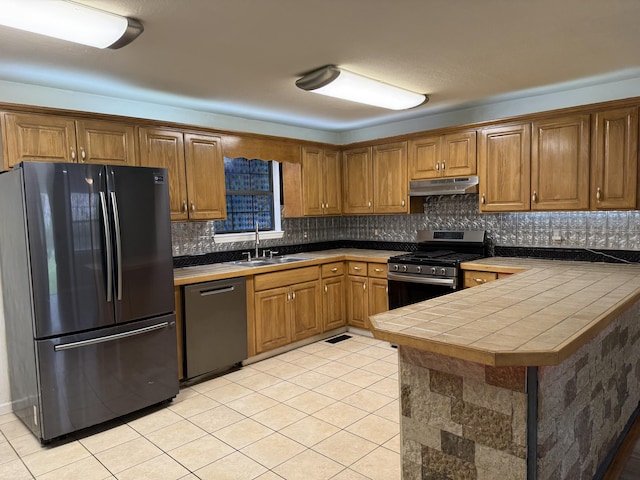 kitchen with a sink, appliances with stainless steel finishes, brown cabinetry, under cabinet range hood, and decorative backsplash