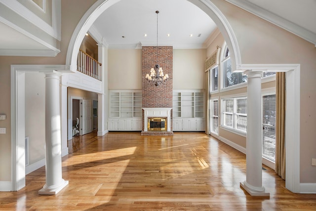 unfurnished living room with a chandelier, light hardwood / wood-style floors, a brick fireplace, ornamental molding, and ornate columns